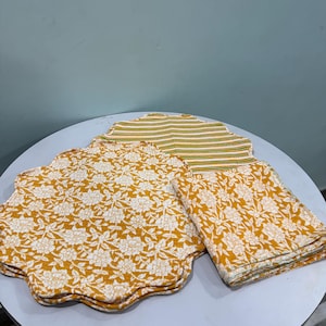 Könnte beinhalten: Ein Set aus Platzdeckchen mit gewelltem Rand und einer gefalteten Serviette aus senfgelbem Stoff mit weißem Blumenmuster. Ein Platzdeckchen zeigt auf der Rückseite ein horizontales Streifenmuster in den Farben Grün, Gelb und Weiß.
