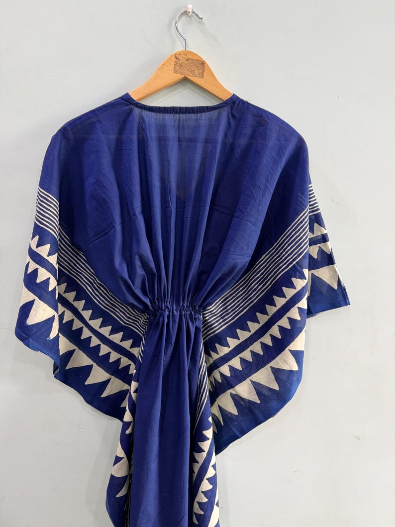 May include: A deep blue kaftan featuring a white geometric pattern along the edges and sleeves. The garment has a cinched waist with a drawstring detail, a V-neckline, and wide, flowing sleeves that drape from the shoulders in a loose silhouette.