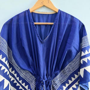 May include: A royal blue kaftan featuring a white geometric pattern along the wide sleeves and hem. The garment has a V-neckline, a drawstring waist with ties, and is displayed on a wooden hanger. The fabric has a visible texture and drape.