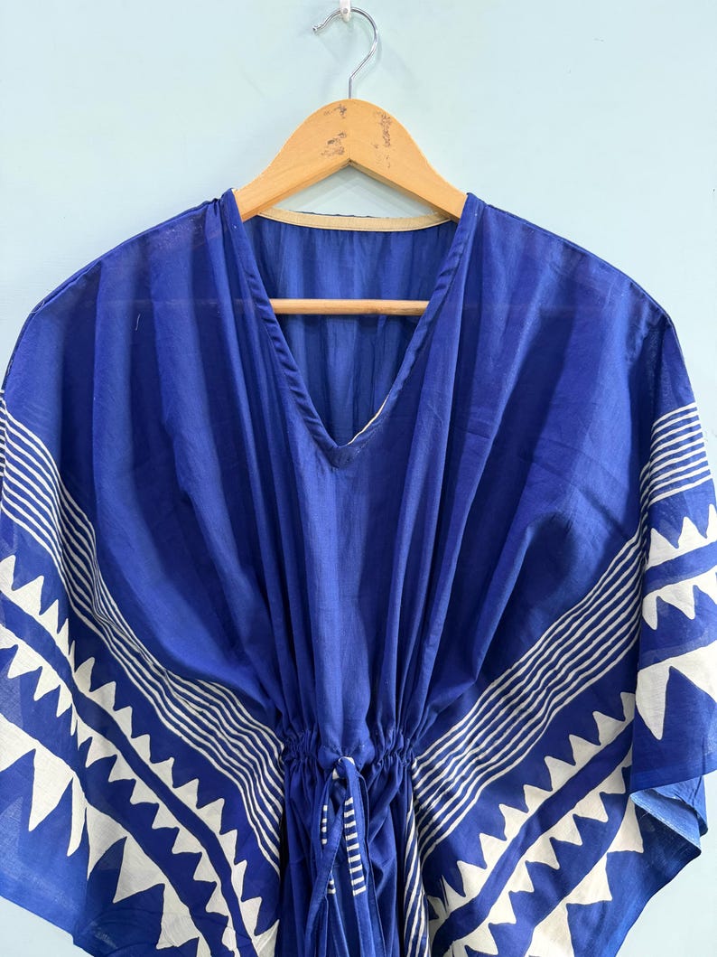 May include: A royal blue kaftan featuring a white geometric pattern along the wide sleeves and hem. The garment has a V-neckline, a drawstring waist with ties, and is displayed on a wooden hanger. The fabric has a visible texture and drape.