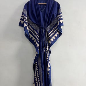 May include: A floor-length kaftan dress in deep blue featuring a white geometric triangle pattern along the edges and sleeves. The garment includes a V-neckline and a central drawstring waist tie that gathers the fabric for a draped silhouette.