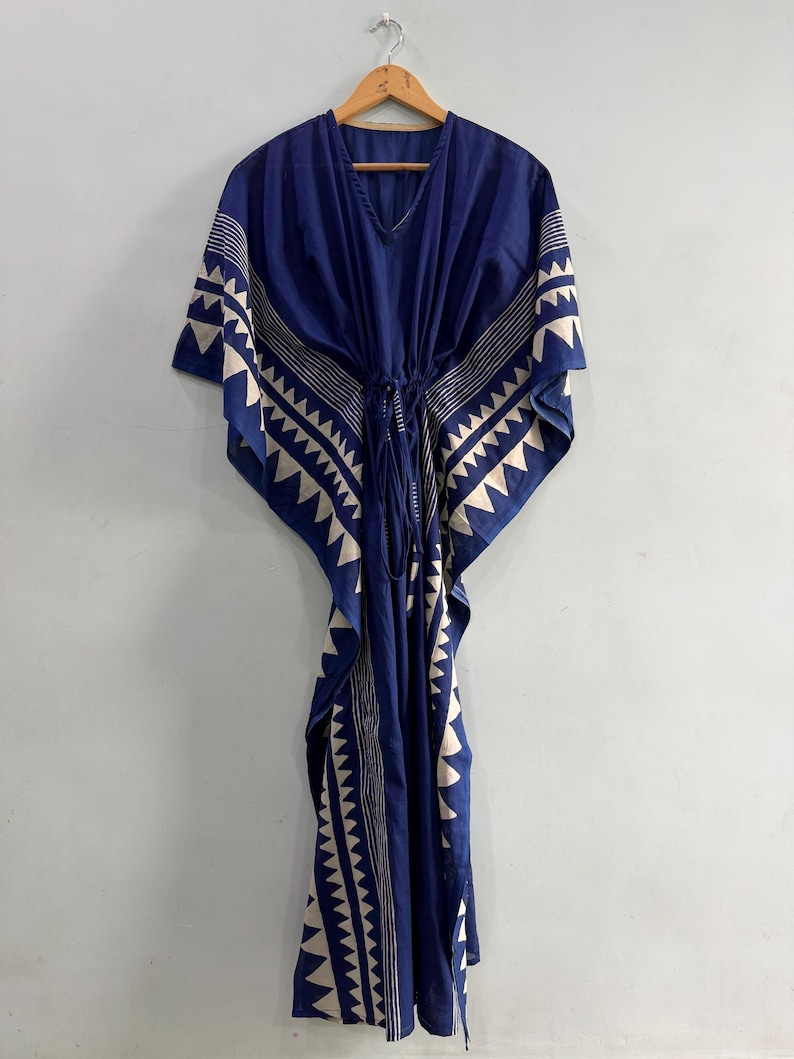 May include: A floor-length kaftan dress in deep blue featuring a white geometric triangle pattern along the edges and sleeves. The garment includes a V-neckline and a central drawstring waist tie that gathers the fabric for a draped silhouette.