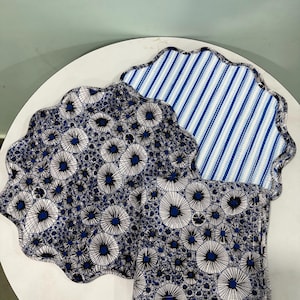 Könnte beinhalten: Zwei Stoff-Platzsets mit gewelltem Rand und eine passende quadratische Serviette. Ein Platzset zeigt ein blau-schwarz-weißes, löwenzahnartiges Blumenmuster, während das andere ein blau-weißes vertikales Streifendesign mit dunkelblauem Rand aufweist.