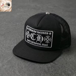 Puede incluir: Gorra de camionero negra con parte trasera de malla y visera curva. La parte delantera presenta un parche rectangular blanco con el texto "Chrome Hearts Hollywood, USA" y un logotipo "CH" estilizado.
