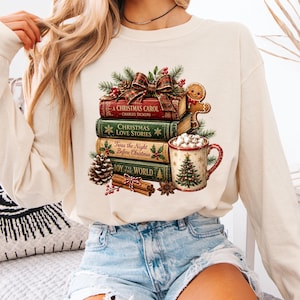 May include: An ivory long-sleeve sweatshirt featuring a graphic print of a stack of four books titled A Christmas Carol, Christmas Love Stories, Twas the Night Before Christmas, and Joy to the World, topped with a bow, pine, and a gingerbread man.