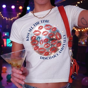 May include: A white short-sleeved t-shirt featuring a graphic of a disco ball covered in red lip prints. The text "KISS ALL THE TIME" is printed in an arc above the disco ball, and "DISCO OCCASIONALLY" is printed in an arc below the disco ball.