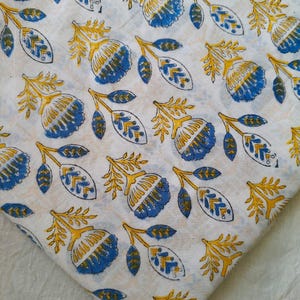 May include: A white cotton fabric featuring a repeating block print pattern of stylised blue flowers with yellow stems and leaves. The floral design is arranged in a scattered layout across the textile, showcasing a hand-stamped aesthetic.