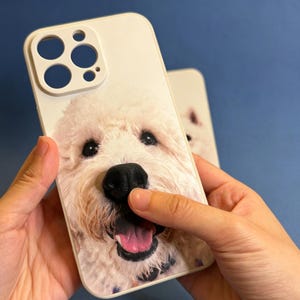 May include: A white smartphone case featuring a high-resolution, close-up photographic print of a fluffy, cream-colored dog with a black nose and an open mouth showing a pink tongue. The case includes a raised camera cutout section at the top.