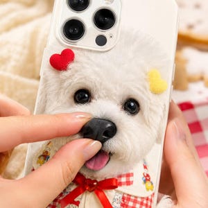 Pode incluir: Uma capa de smartphone branca com o rosto de um cão branco e felpudo, nariz preto, língua rosa e um coração vermelho. O cão usa uma gola creme com um laço vermelho e um padrão de xadrez vermelho e branco na parte inferior da capa.