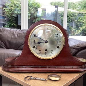 Puede incluir: Un reloj de sobremesa de madera marrón oscuro con la parte superior redondeada y una esfera circular con números arábigos negros, un bisel dorado y manecillas negras. Una llave de cuerda metálica se encuentra sobre la superficie frente al reloj.