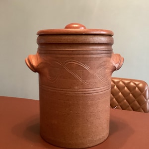 May include: A cylindrical terracotta-colored stoneware crock with a matching lid and two side handles. The exterior features a textured surface with horizontal incised lines and a decorative swirling pattern carved into the upper section.