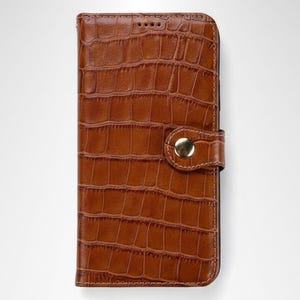 May include: A rectangular brown phone case with a textured crocodile skin pattern. The case features a fold-over flap secured by a single gold-toned metal snap button. The text CORIUMLAB is printed in a dark font at the bottom left corner.
