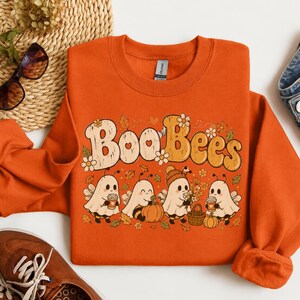 May include: An orange crewneck sweatshirt featuring a graphic print with the text "Boo Bees" in a retro font. The design includes four ghost characters dressed as bees, surrounded by autumn leaves, small flowers, pumpkins, and coffee cups.