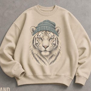 May include: A beige crewneck sweatshirt featuring a central graphic illustration of a white tiger wearing a blue-grey knit beanie. The tiger is depicted from the chest up with detailed fur textures, forward-facing eyes, and a neutral expression.
