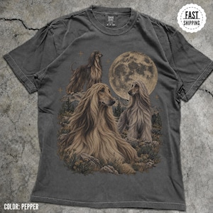 May include: A charcoal grey t-shirt featuring a graphic print of three long-haired Afghan Hound dogs in a mountainous landscape under a large full moon. The shirt has a crew neckline and short sleeves, with a Comfort Colors brand label at the neck.