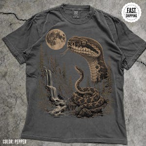 Peut inclure: Un t-shirt couleur poivre avec une impression graphique représentant une grande tête de serpent et un serpent enroulé sur un rocher. La scène comprend une cascade, des pins et une pleine lune en arrière-plan. Col rond et manches courtes.