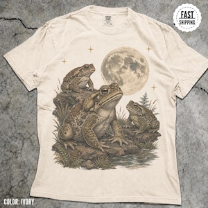 May include: An ivory short-sleeve t-shirt featuring a graphic print of three toads sitting on rocks near a pond under a large full moon. The design includes small star icons and grassy vegetation. The shirt has a crew neckline and a Comfort Colors label.