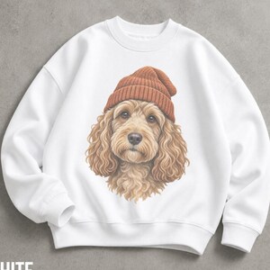 May include: A white long-sleeve crewneck sweatshirt featuring a central graphic illustration of a curly-haired golden-brown dog wearing a ribbed rust-coloured beanie hat. The dog has large dark eyes and a detailed, realistic fur texture.