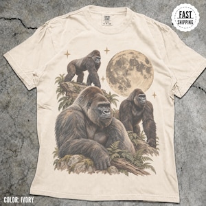 May include: An ivory short-sleeved t-shirt featuring a graphic print of three gorillas in a jungle setting with a large full moon and stars in the background. The shirt has a crew neckline and a Comfort Colors brand label at the inner neck.