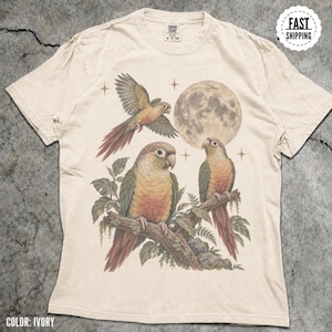 May include: An ivory short-sleeve t-shirt featuring a graphic print of three green and orange parrots. One parrot is in flight, while two others are perched on a branch. A large full moon and four-pointed stars are visible in the background.