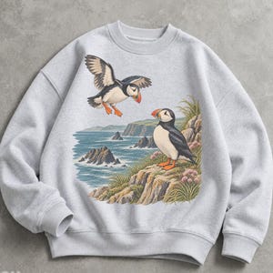 May include: A light gray crewneck sweatshirt featuring a graphic print of two Atlantic puffins. One puffin is in flight while the other stands on a rocky cliff overlooking a blue ocean with jagged sea stacks and green coastal vegetation.