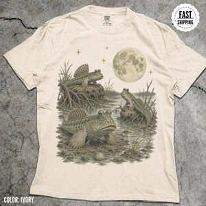 May include: An ivory-colored short-sleeve t-shirt featuring a detailed graphic illustration of three mudskipper fish in a marshy habitat under a full moon and stars. The design is rendered in shades of brown and green, showcasing the fish on mud.