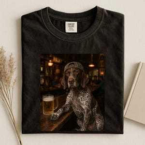May include: A black crew neck t-shirt featuring a graphic print of a brown and white spotted dog wearing a baseball cap, sitting at a wooden bar next to a glass of beer. The shirt has a white interior neck label with the text Comfort Colors.