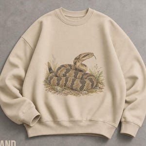 May include: A beige crewneck sweatshirt featuring a detailed graphic illustration of a coiled rattlesnake with brown and tan scales, positioned amidst small patches of grass and rocks in the centre of the chest area of the garment.