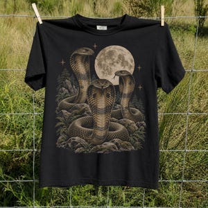 May include: A black short-sleeve t-shirt featuring a graphic print of three cobras coiled on rocky terrain. The design includes a large full moon in the background, surrounded by pine trees and small four-pointed stars in a muted beige color palette.