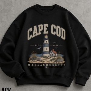 May include: A black crewneck sweatshirt featuring a graphic print of a lighthouse on a sandy dune with a small house, beach grass, and birds. The text reads CAPE COD, EST. 1602, and MASSACHUSETTS in a vintage-style font with a compass rose icon.
