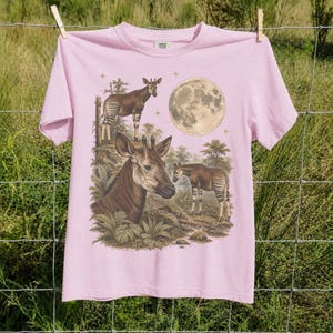 May include: A light pink short-sleeve t-shirt featuring a graphic print of three okapis in a forest setting with a large full moon and small stars. The shirt has a crew neckline and a label at the inner neck area with the text Comfort Colors.