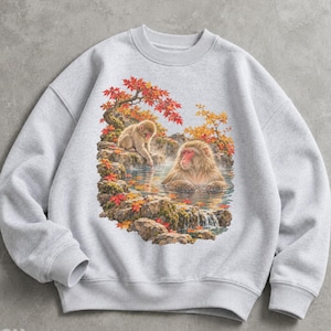 May include: A light grey crewneck sweatshirt featuring a graphic print of two Japanese macaques in a hot spring surrounded by rocky terrain and autumn foliage with vibrant red and orange maple leaves against a natural landscape.