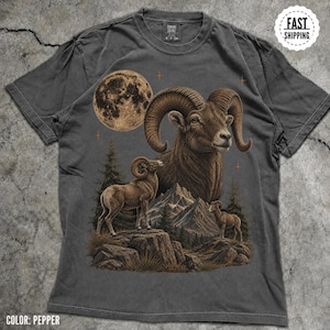 Peut inclure: Un t-shirt gris anthracite avec une impression graphique représentant une grande tête de mouflon, deux mouflons plus petits sur un terrain rocheux, une pleine lune, des pins et des sommets montagneux dans des tons marron et beige.