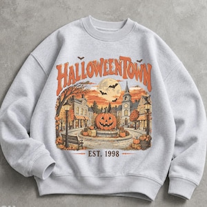 May include: A light gray crewneck sweatshirt featuring a graphic print of a town square at sunset. The text HALLOWEENTOWN is arched above a large jack-o'-lantern, buildings, street lamps, bats, and a full moon, with EST. 1998 printed below.