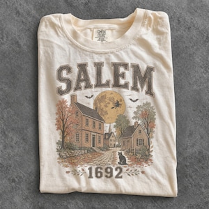 May include: A cream-coloured t-shirt featuring a graphic print of a colonial town scene with the text SALEM at the top and 1692 at the bottom. The illustration includes houses, a full moon with a witch silhouette, bats, and a black cat.