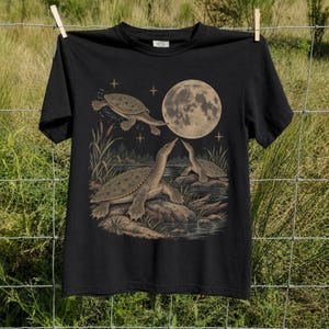 May include: A black short-sleeved t-shirt featuring a graphic print of three turtles in a pond setting under a large full moon. The illustration includes bulrushes, rocks, and small four-pointed stars in muted beige and brown tones against the black fabric.