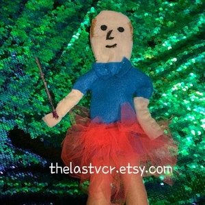 May include: A handmade felt doll with a blue shirt and a pink tulle skirt. The doll is holding a stick in its hand. The doll is standing on a green sparkly background. Thelastvcr.etsy.com is written on the background.