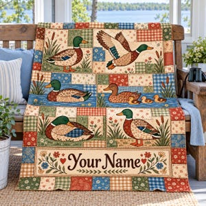 May include: A patchwork quilt featuring mallard ducks in various poses, including flying and swimming with ducklings. The design incorporates floral and plaid patterns in green, blue, red, and tan. The bottom panel displays the text Your Name.