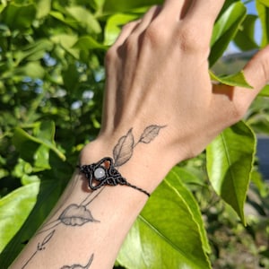 Può includere: Un braccialetto in macramè nero con un design centrale che tiene una piccola pietra rotonda di colore chiaro. Il braccialetto è indossato su un polso con tatuaggi di foglie, su uno sfondo di fogliame verde.