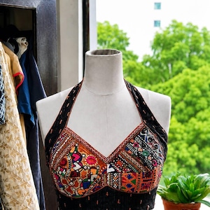 May include: A black halter top featuring a vibrant, multi-colored patchwork design across the chest. The garment is adorned with intricate embroidery and small mirror-like accents, showcasing a variety of geometric patterns in orange, red, blue, and green.