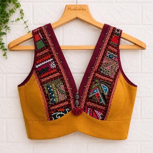 May include: Mustard yellow sleeveless top with a deep V-neck. The top features embroidered panels in shades of red, green, blue, and pink. A small tassel and decorative buttons adorn the centre. The top is hanging on a wooden hanger.
