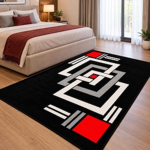 May include: A rectangular area rug featuring a black background with a central geometric pattern composed of interlocking white and grey squares, accented by solid red square blocks and parallel white and grey lines at the ends of the design.