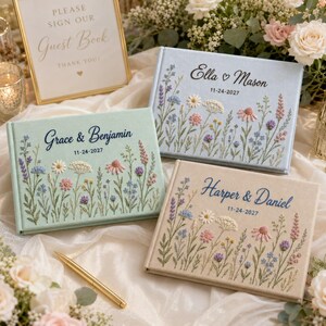Könnte beinhalten: Drei rechteckige Gästebücher mit Stoffeinbänden in Hellblau, Salbeigrün und Beige. Jedes Buch ist mit gestickten Wildblumen, personalisierten Namen und einem Datum versehen. Ein goldener Stift und ein Schild mit der Aufschrift Please Sign Our Guest Book.
