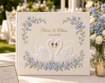 Puede incluir: Un libro de visitas de color crema con un diseño bordado de dos cisnes blancos frente a frente, rodeados de enredaderas florales azules. El texto "Olivia & Ethan" y "09.21.2026" está bordado en hilo dorado en el centro de la portada.