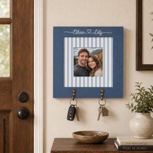 Könnte beinhalten: Ein quadratischer, blauer Schlüsselhalter aus Holz zur Wandmontage mit einem zentralen Fotorahmen auf hellblau-weiß gestreiftem Hintergrund. Der Rahmen ist mit dem weißen Schriftzug Ethan und Lily und einem Herzsymbol personalisiert. Zwei Metallhaken halten Schlüssel.