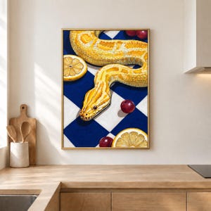May include: A framed wall art print featuring a yellow snake winding across a blue and white chequered background. The composition includes scattered red cherries and two sliced lemon halves, all rendered in a detailed, illustrative style.