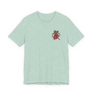 May include: A light mint green short-sleeve t-shirt with a crew neckline. On the upper left chest, there is a small embroidered graphic of a red strawberry wearing a brown hat with a green brim and a pink ribbon tied around the stem.