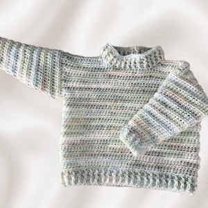 Könnte beinhalten: Ein langärmeliger Häkelpullover mit einem melierten Garnmuster in sanften Pastelltönen aus Blau, Grün, Rosa und Weiß. Das Kleidungsstück hat einen gerippten Stehkragen, gerippte Bündchen und einen gerippten Saum, mit dem Text Kimis Stoffliebe in der Ecke.