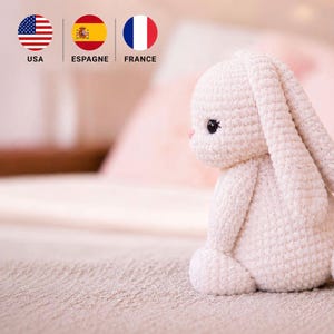 Peut inclure: Un lapin en peluche crocheté de couleur crème avec de longues oreilles tombantes et de petits yeux noirs. L'animal est fabriqué en fil avec un motif de points texturé visible, présentant une tête et un corps arrondis en position assise.