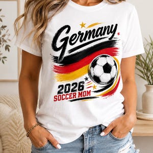 Op de afbeelding: Een wit t-shirt met korte mouwen en een grafische print met het woord Germany in zwart handschrift, een voetbal en de tekst 2026 SOCCER MOM in zwart en rood. Het ontwerp bevat penseelstreekaccenten in de kleuren zwart, rood en geel.